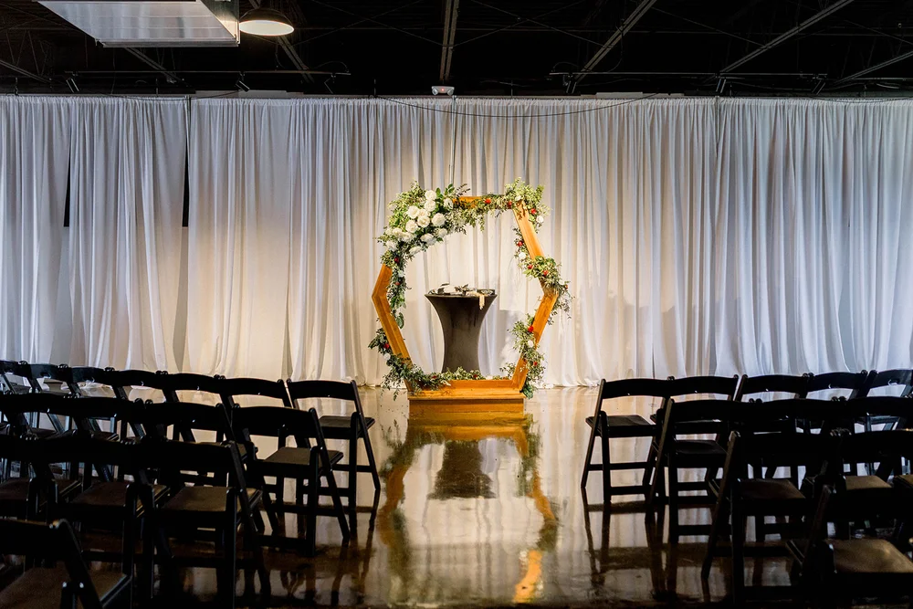 Wedding ceremony set up at Radius Indianapolis with white draping, a wooden hexagon arch with florals and greenery, and black folding chairs.