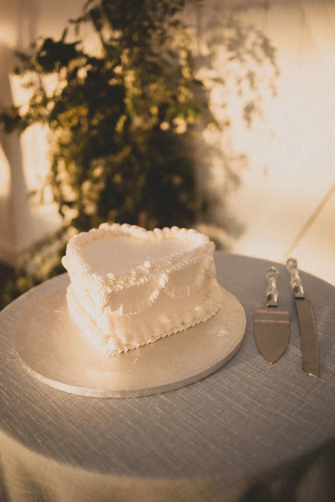 A white frosted heart-shaped wedding cake on a silver platter with silver cake cutting tools on the table next to it.