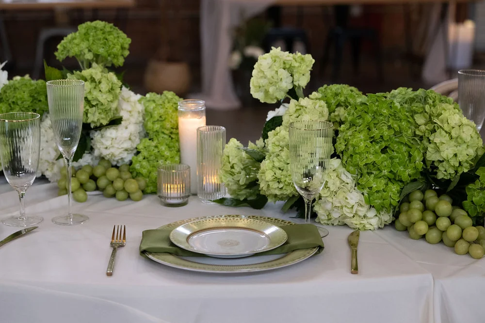 Table setting with lush green florals and fruit, green plates and napkins, and gold cutlery set by wedding planner.