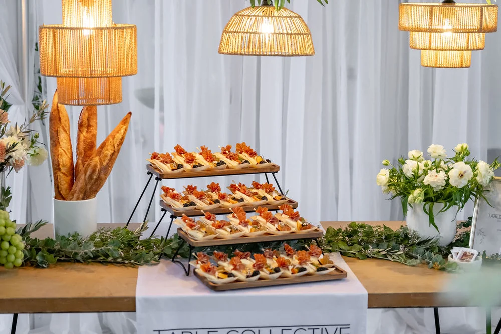 Appetizer display with a tiered shelving unit for small bites, a container of large loaves of bread, and florals set by event planner.