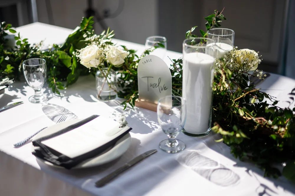 Classic wedding table with white linens and plates, candles, greenery and white roses as the centerpiece at Indianapolis wedding venue.