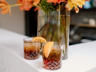 bourbon-old-fashioned-cocktails-bar-drinks-828-Venues Two Old Fashioned cocktails with orange garnishes on a white bar top, with a vase of flowers visible in the background.