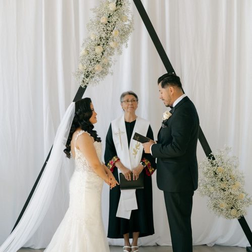 A bride and groom holding hands and exchanging vows during their indoor wedding ceremony, standing under a modern black geometric arch decorated with white flowers.