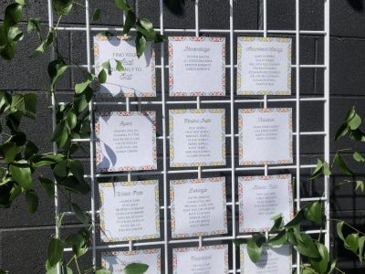wedding-seating-chart-grid-floral-decor-828-Venues A wedding seating chart displayed on a white grid with green vines, showing guest names and table numbers on individual cards with a floral pattern.